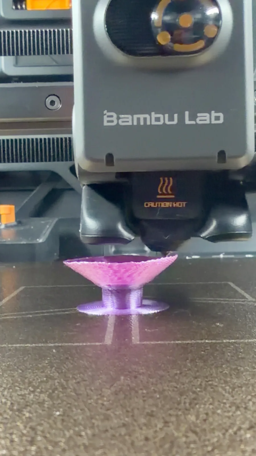 Печатающая головка Bambu Lab крупным планом: сопло печатает фиолетовый образец основания на столе